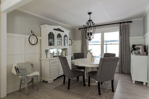 Uma sala de jantar com uma mesa branca, cadeiras cinzas, um armário branco com portas de vidro e um candelabro.