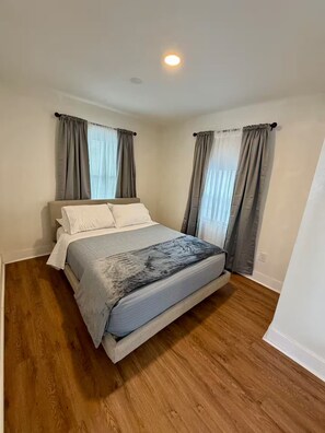 A bedroom with a bed, wooden flooring, and two windows with curtains. - Golden safari home Perfect Getaway for Relaxation (Tampa)