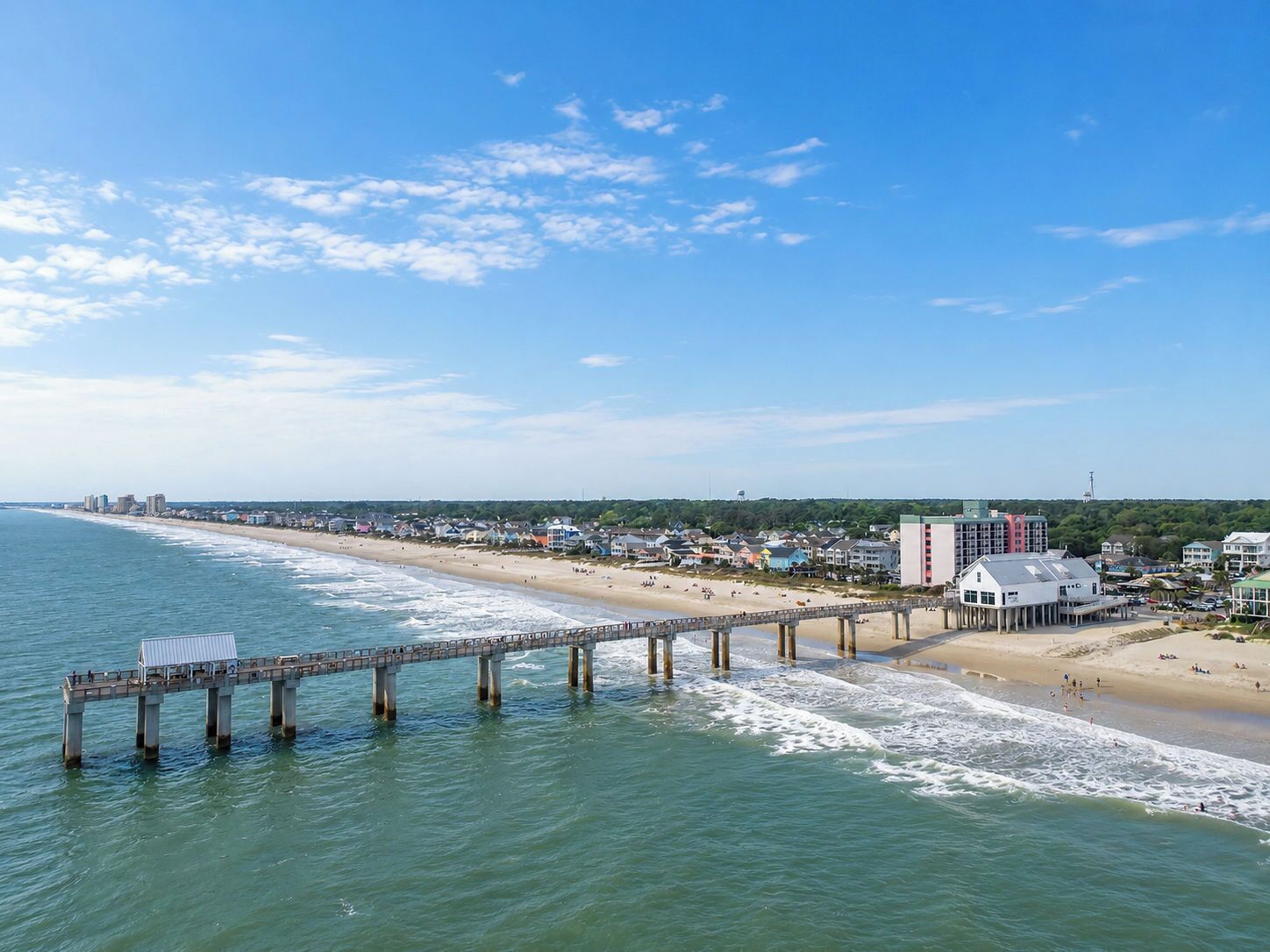 A coastal cityscape with a long pier extending into the sea, a beach, and residential buildings.