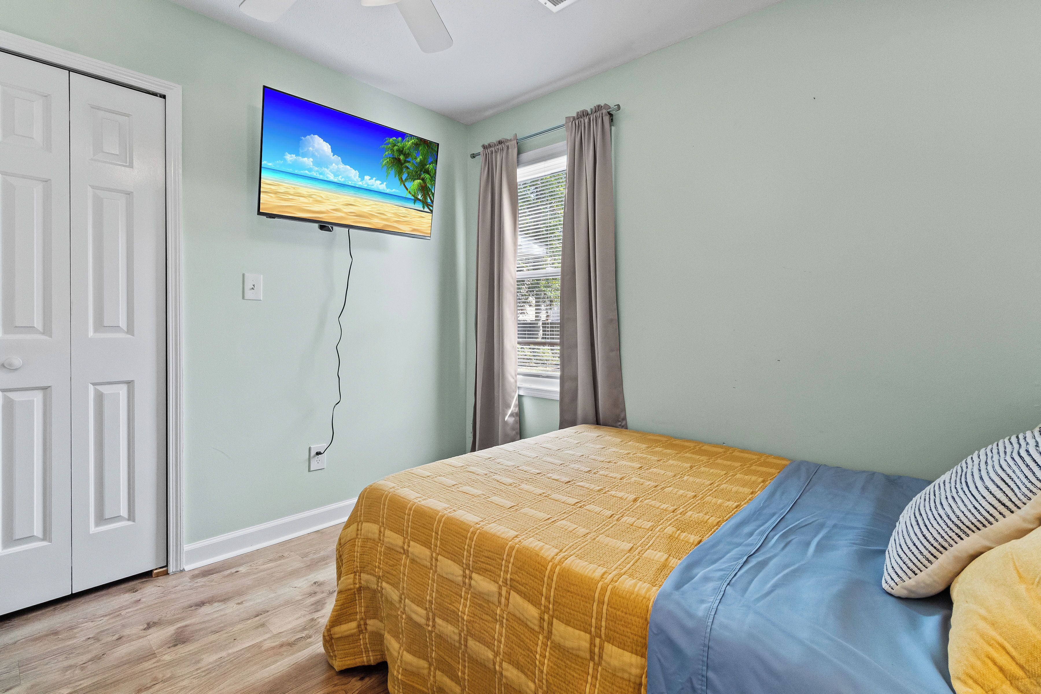 Une chambre à coucher avec un lit, un téléviseur fixé au mur, une fenêtre avec des rideaux et une porte.