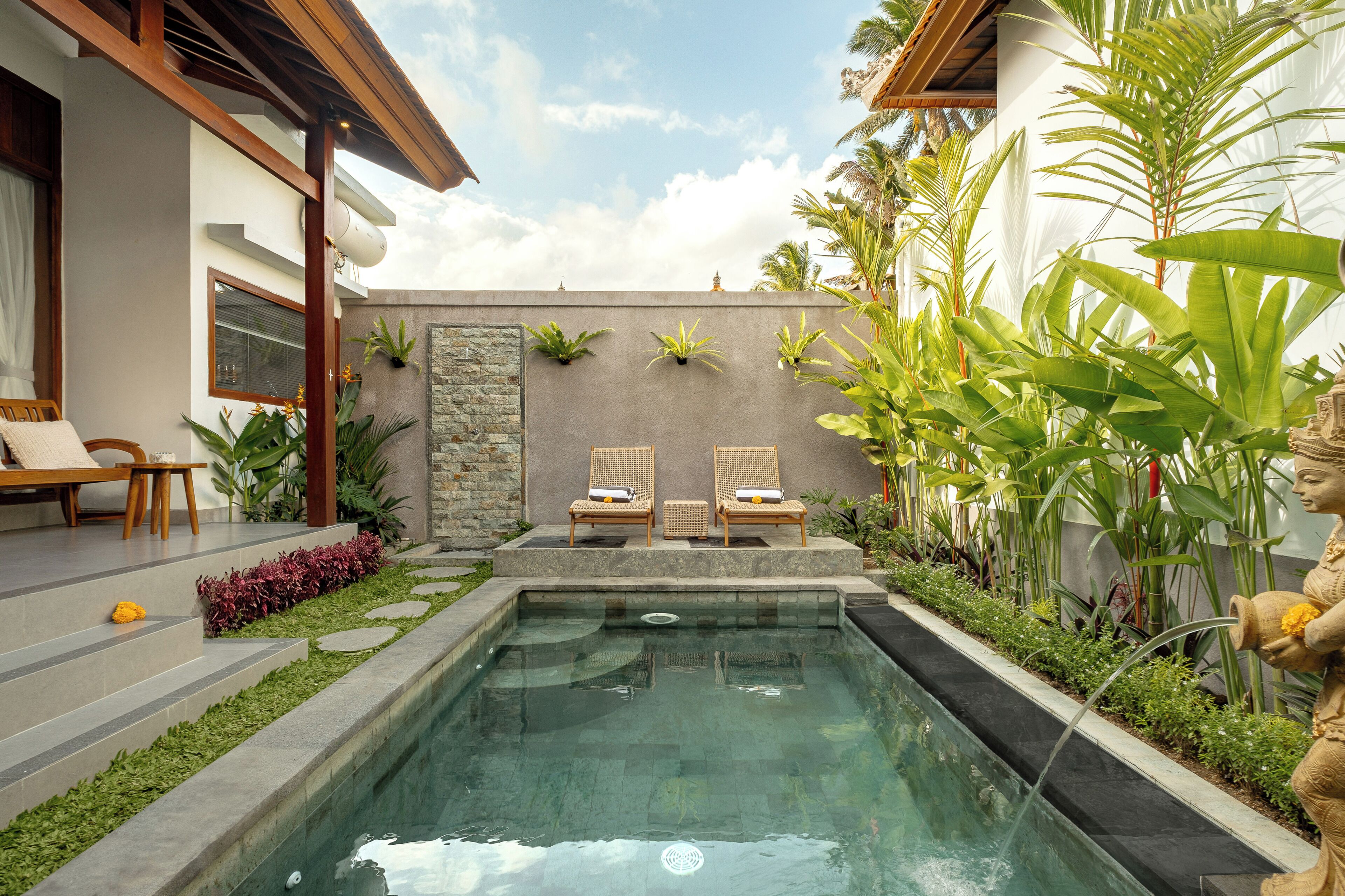 A swimming pool with two lounge chairs, surrounded by tropical plants and a stone wall.