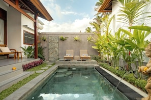 Piscina con dos sillas de descanso, rodeada de plantas tropicales y un muro de piedra.