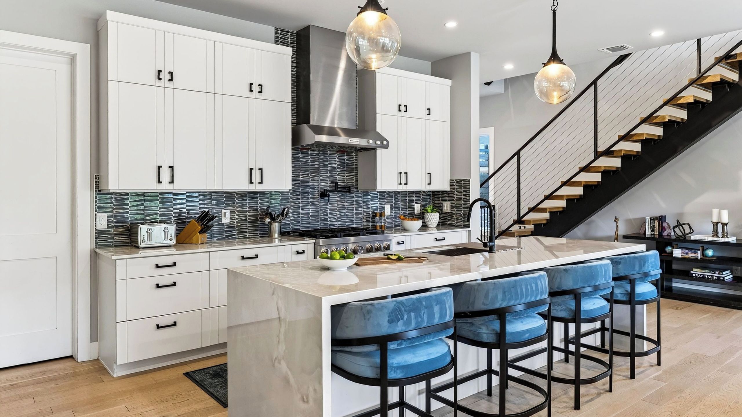 Una cocina moderna con armarios blancos, una isla central con taburetes y una escalera que lleva a otro nivel.