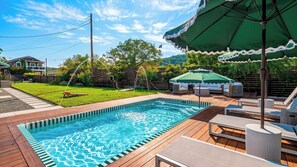 Une piscine avec une terrasse en bois, du mobilier d’extérieur et un parasol vert.
