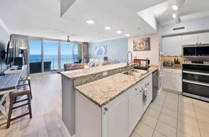 A modern kitchen with a granite countertop, white cabinets, and a dining area with a table and chairs. - Emerald Beach - A Gulf Front Resort Experience (Panama City Beach)