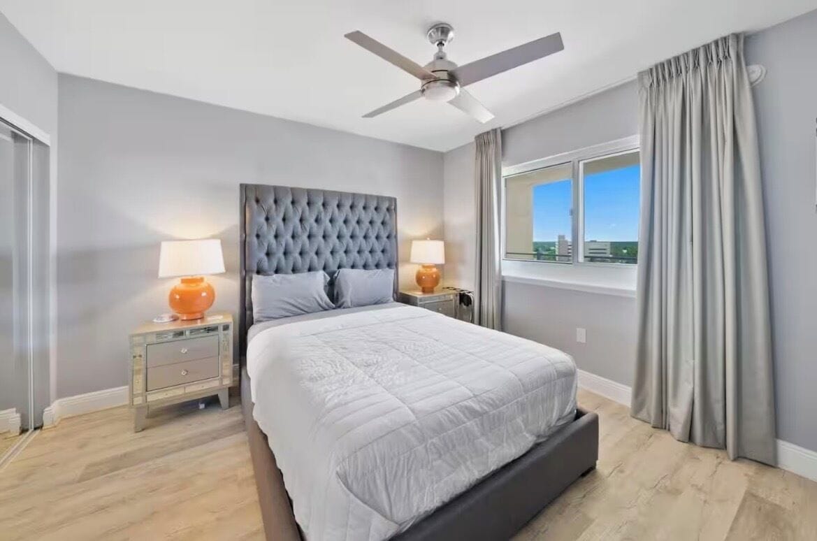 Une chambre à coucher avec un grand lit, deux tables de chevet avec des lampes, un ventilateur de plafond et une fenêtre avec des rideaux.