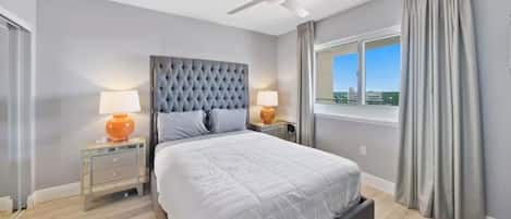 Une chambre à coucher avec un grand lit, deux tables de chevet avec des lampes, un ventilateur de plafond et une fenêtre avec des rideaux.
