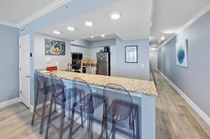 A modern kitchen with a bar area, clear bar stools, and a refrigerator. - Emerald Beach - A Gulf Front Resort Experience (Panama City Beach)