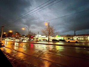 Eine nasse Straße bei Nacht mit parkenden Autos am Straßenrand und beleuchteten Geschäften im Hintergrund.