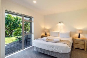 A bedroom with a large bed, two bedside tables, a window with a view of a garden, and a sliding glass door. - Views on Beachman - pet friendly with lake views (Taupo)