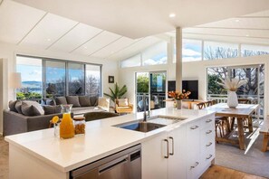 A modern kitchen with a large island, a dining area with a wooden table, and a living room with a sofa and a TV. - Views on Beachman - pet friendly with lake views (Taupo)