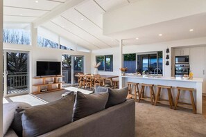 A modern open-plan living area with a sofa, TV, dining table, and kitchen counter. - Views on Beachman - pet friendly with lake views (Taupo)