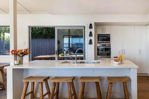 A modern kitchen with a central island, bar stools, a built-in oven, and a view through a glass door. - Views on Beachman - pet friendly with lake views (Taupo)