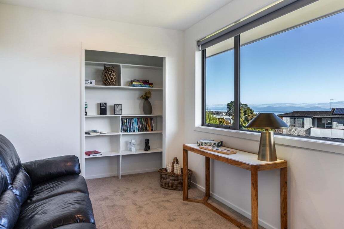 Ein modernes Wohnzimmer mit einem Ledersofa, einem weißen Bücherregal, einem Holztisch und Blick durch ein großes Fenster.