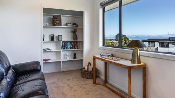 A modern living room with a leather sofa, a white bookshelf, a wooden side table, and a view through a large window. - Views on Beachman - pet friendly with lake views (Taupo)