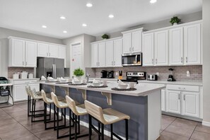 A modern kitchen with white cabinetry, a central island with bar stools, a refrigerator, a microwave, and a coffee maker. - 7BR Solara Resort Home with Private Pool & Games (Kissimmee)