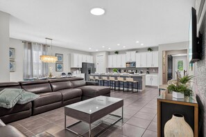 A modern kitchen with white cabinets, a central island, and a dining area with a brown leather sofa and a glass coffee table. - 7BR Solara Resort Home with Private Pool & Games (Kissimmee)