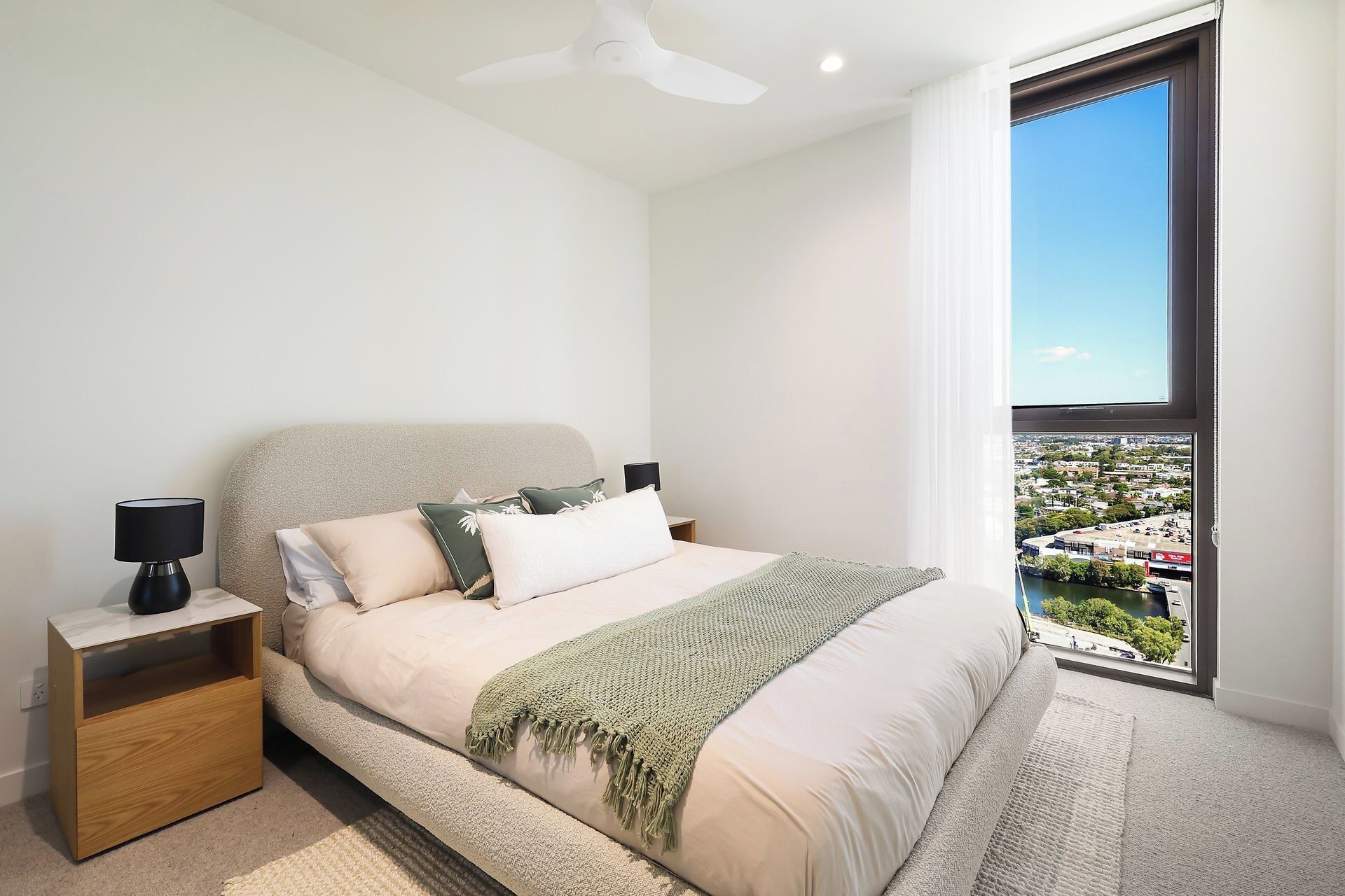 A modern bedroom with a bed, nightstand, and a view of a cityscape through a large window.