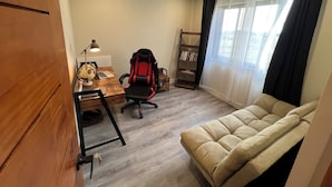 A room with a wooden desk, a laptop, a red and black gaming chair, a bookshelf, a beige sofa, and a window with sheer curtains.