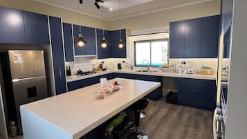 A modern kitchen with a central island, dark blue cabinets, stainless steel appliances, pendant lighting, and a window providing natural light.