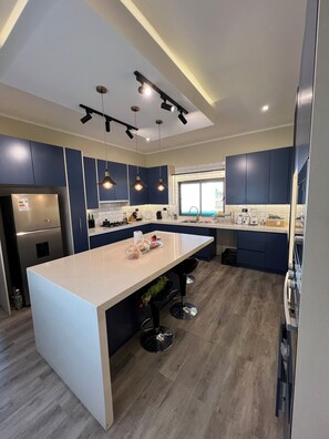 A modern kitchen with a central island, dark blue cabinets, stainless steel appliances, pendant lighting, and a window providing natural light.