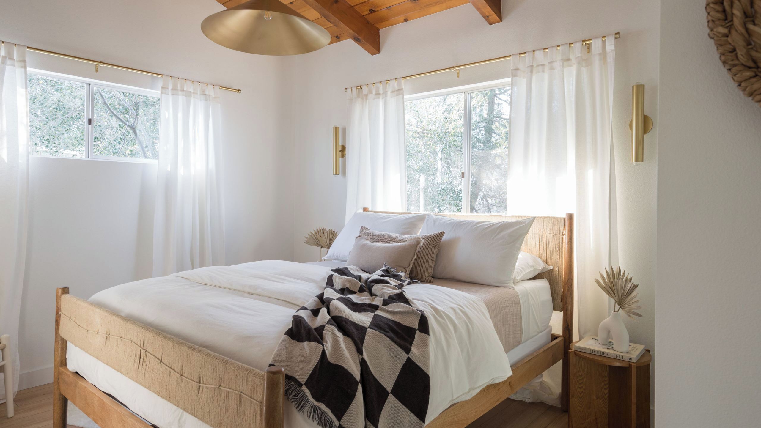 Une chambre avec un lit en bois, une literie blanche et un plaid à carreaux. On y trouve deux tables de chevet ornées de vases décoratifs et un livre intitulé « VIVEZ PLEINEMENT ».