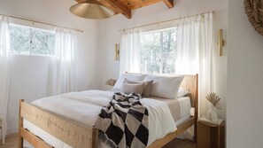 Un dormitorio con cama de madera, ropa de cama blanca y una manta de cuadros. Hay dos mesitas de noche con jarrones decorativos y un libro titulado 'LIVE BEAUTIFUL'.
