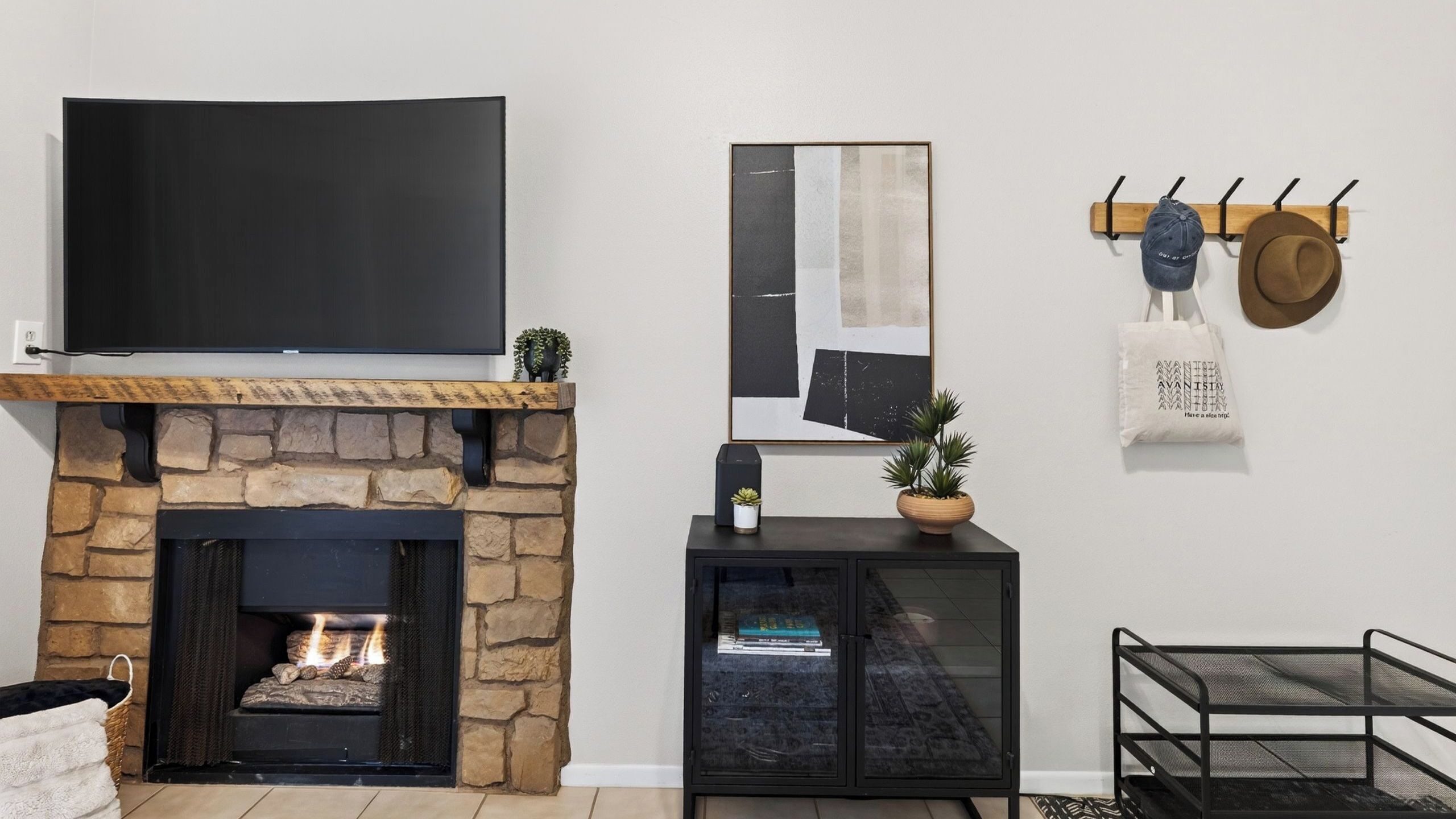 A living room with a stone fireplace, a flat-screen TV mounted above it, a modern art piece, a coat rack with hats, and a small cabinet with a plant.