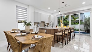 Une cuisine moderne avec des meubles blancs, un coin repas avec une table en bois et des chaises en osier, et une vue sur l’extérieur grâce à de grandes fenêtres.
