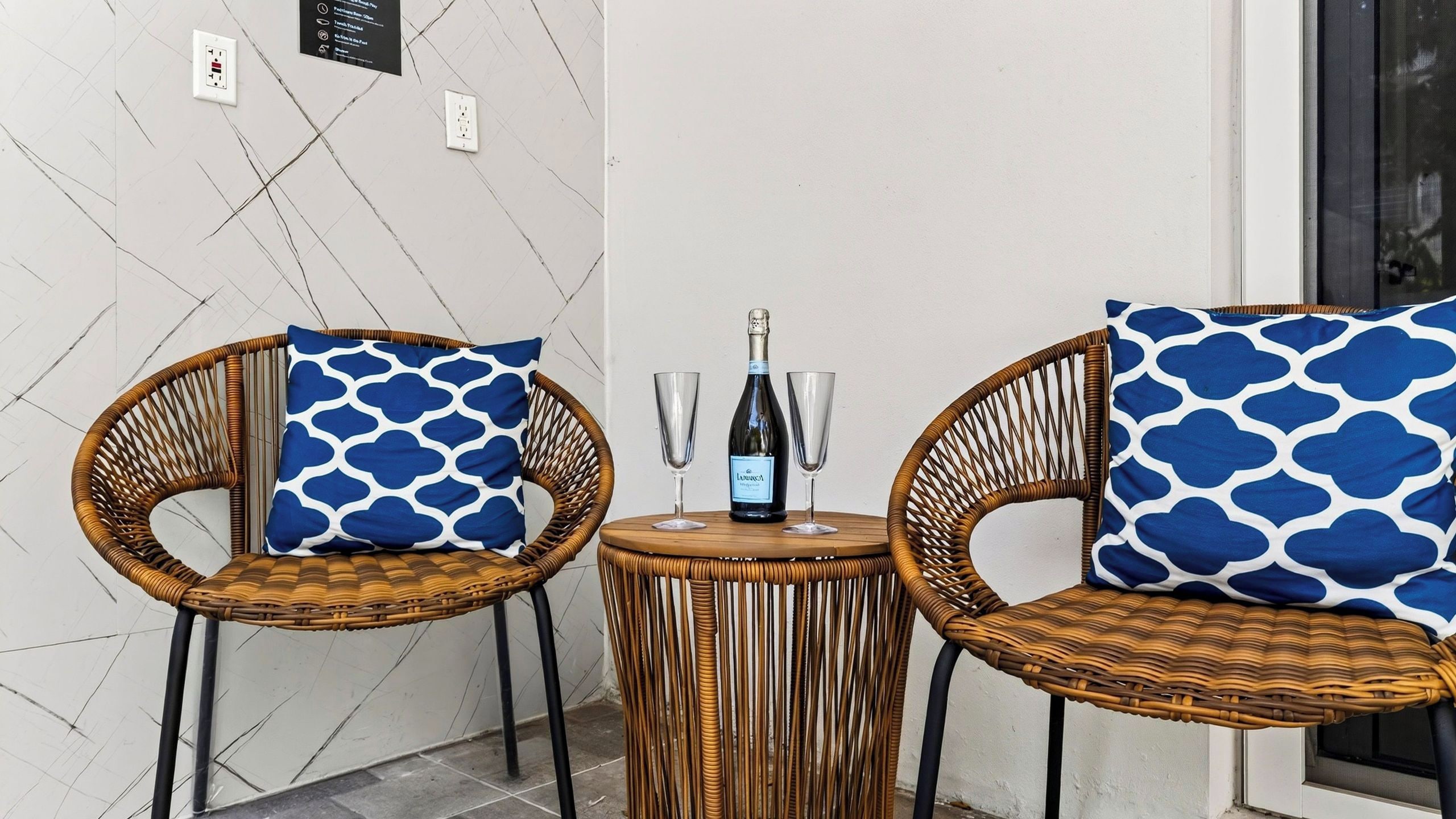 Two wicker chairs with blue patterned cushions, a small round table with a bottle of wine and two glasses, and a wall with electrical outlets and a sign.