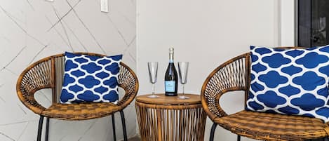 Deux chaises en osier avec des coussins à motifs bleus, une petite table ronde avec une bouteille de vin et deux verres, et un mur équipé de prises électriques et d’un panneau indicateur.