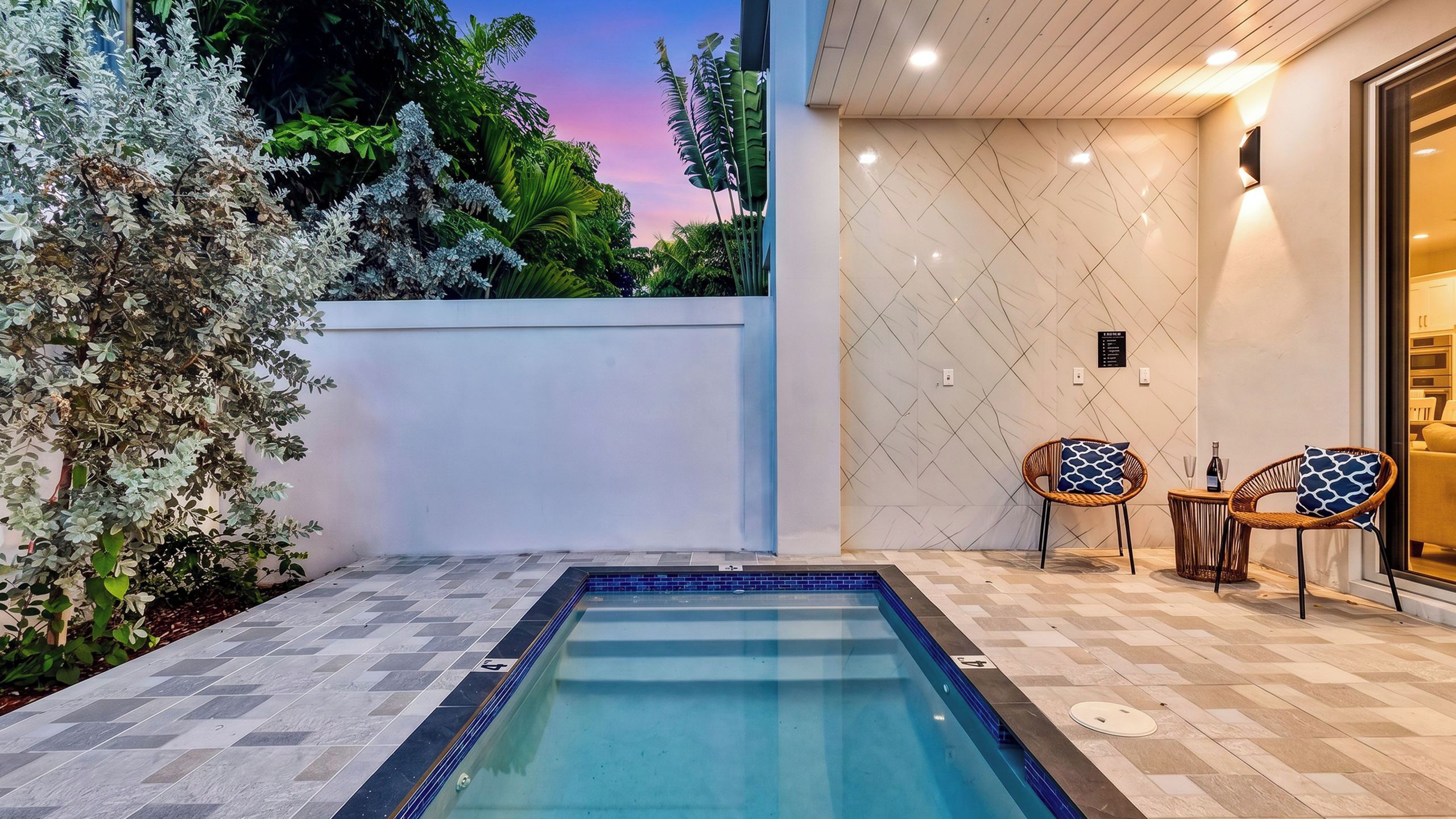 A swimming pool with a tiled deck, two wicker chairs with patterned cushions, and a wall with a geometric tile pattern.