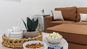 Ein Wohnzimmer mit einem braunen Sofa, einer weißen Schüssel mit Popcorn, einer weißen Schüssel mit Brezeln, einem weißen Dekorationsgegenstand und einer Topfpflanze.