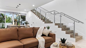 Un salon moderne avec un canapé en cuir marron, un escalier en verre avec des marches en bois et une vue sur l’extérieur grâce à de grandes fenêtres.