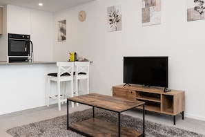 A modern living room with a TV, wooden entertainment unit, white chairs, and a small table.