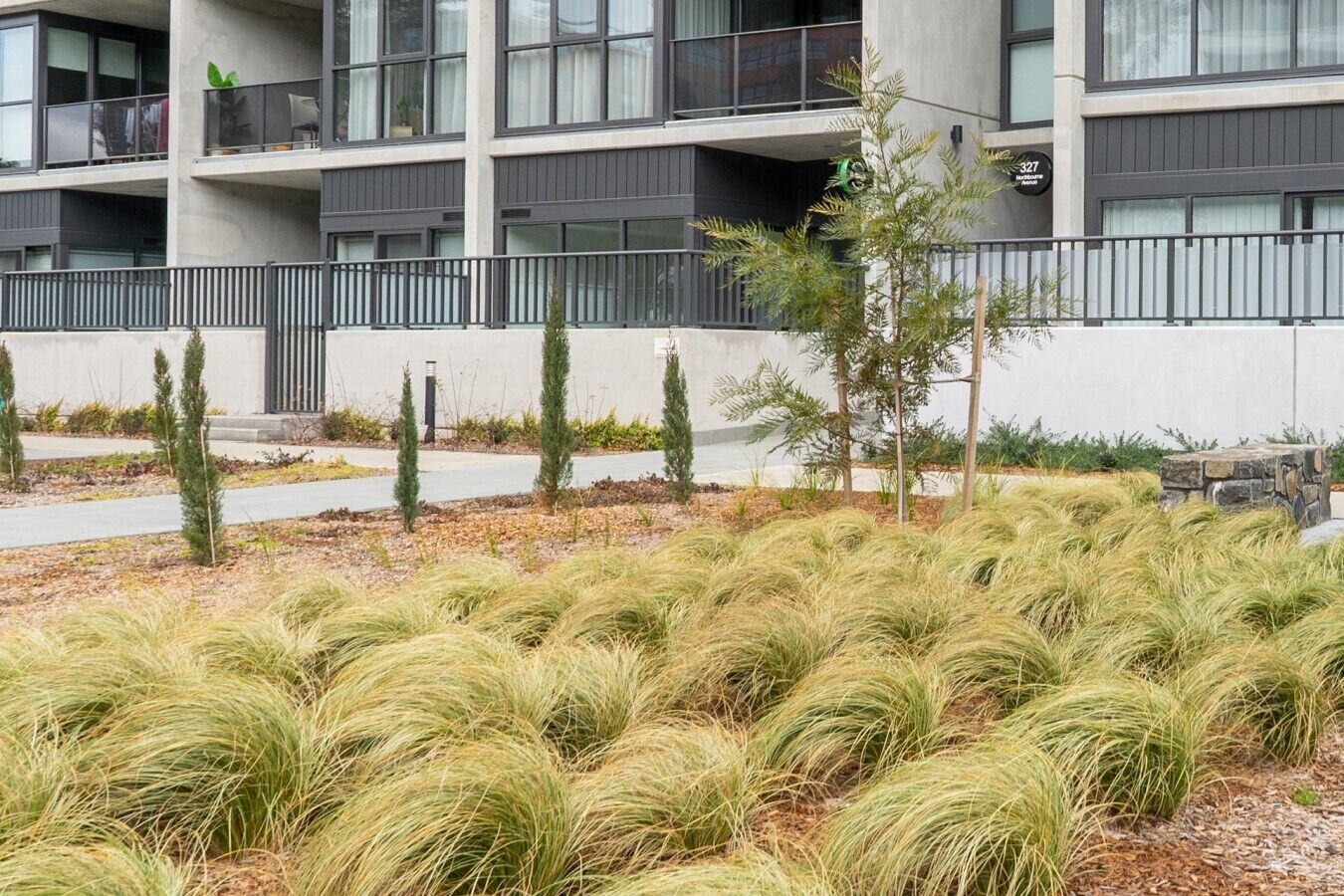 A modern building with a balcony, a stone wall, and a grassy area with tall grasses.