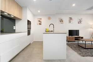 A modern kitchen with white cabinetry, a central island with a sink, and a flat-screen TV on a wooden stand. - Inner North 1BR Near Braddon & Dickson (Lyneham)
