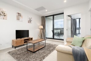 A modern living room with a flat-screen TV, wooden furniture, a glass door leading to an outdoor balcony, and a sofa with a throw blanket. - Inner North 1BR Near Braddon & Dickson (Lyneham)