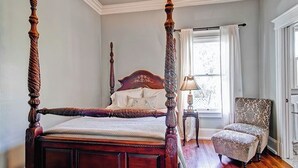 A four-poster bed with a wooden headboard and footboard, a nightstand with a lamp, a patterned armchair, and a window with curtains.