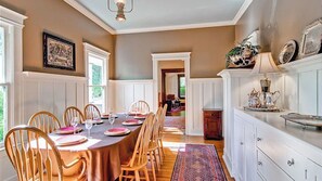 Ein Esszimmer mit einem langen Tisch, gedeckt für eine Mahlzeit, Holzsesseln und einer weißen Kommode mit Dekorationen.