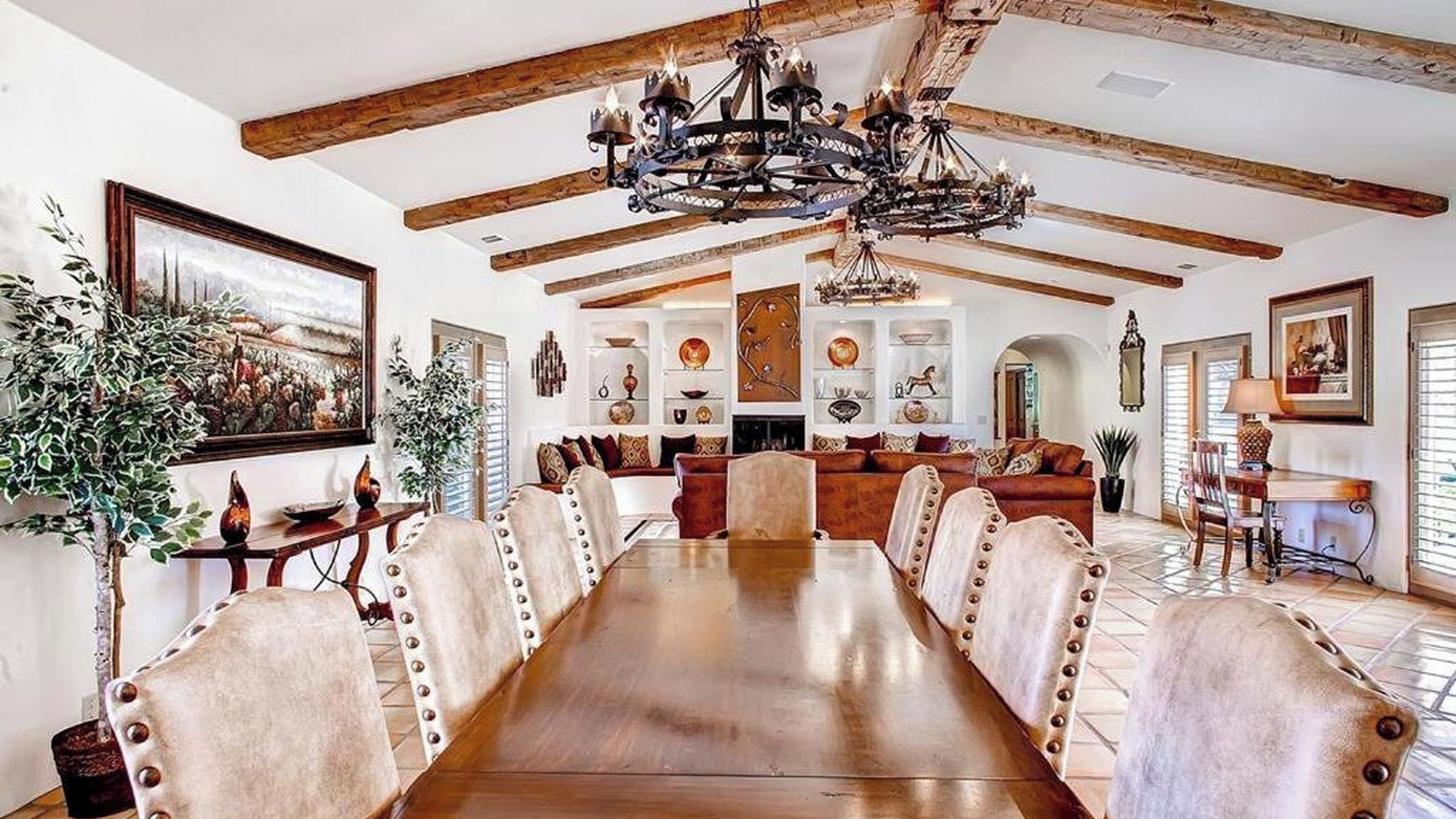 A spacious dining area with a long wooden table, upholstered chairs with rivets, and a decorative chandelier.