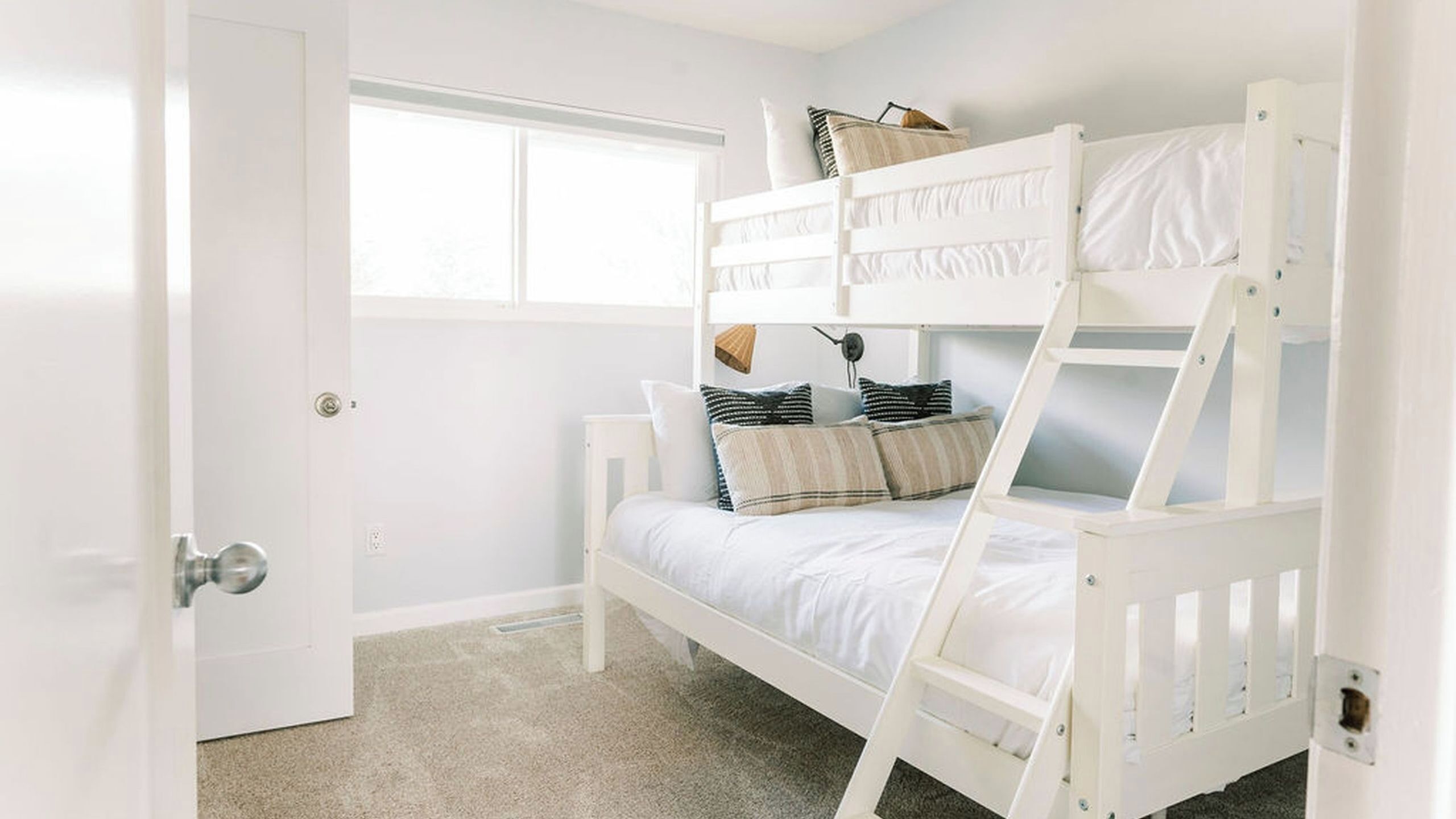 A bunk bed with white bedding and pillows, a window with blinds, and a door with a silver handle.