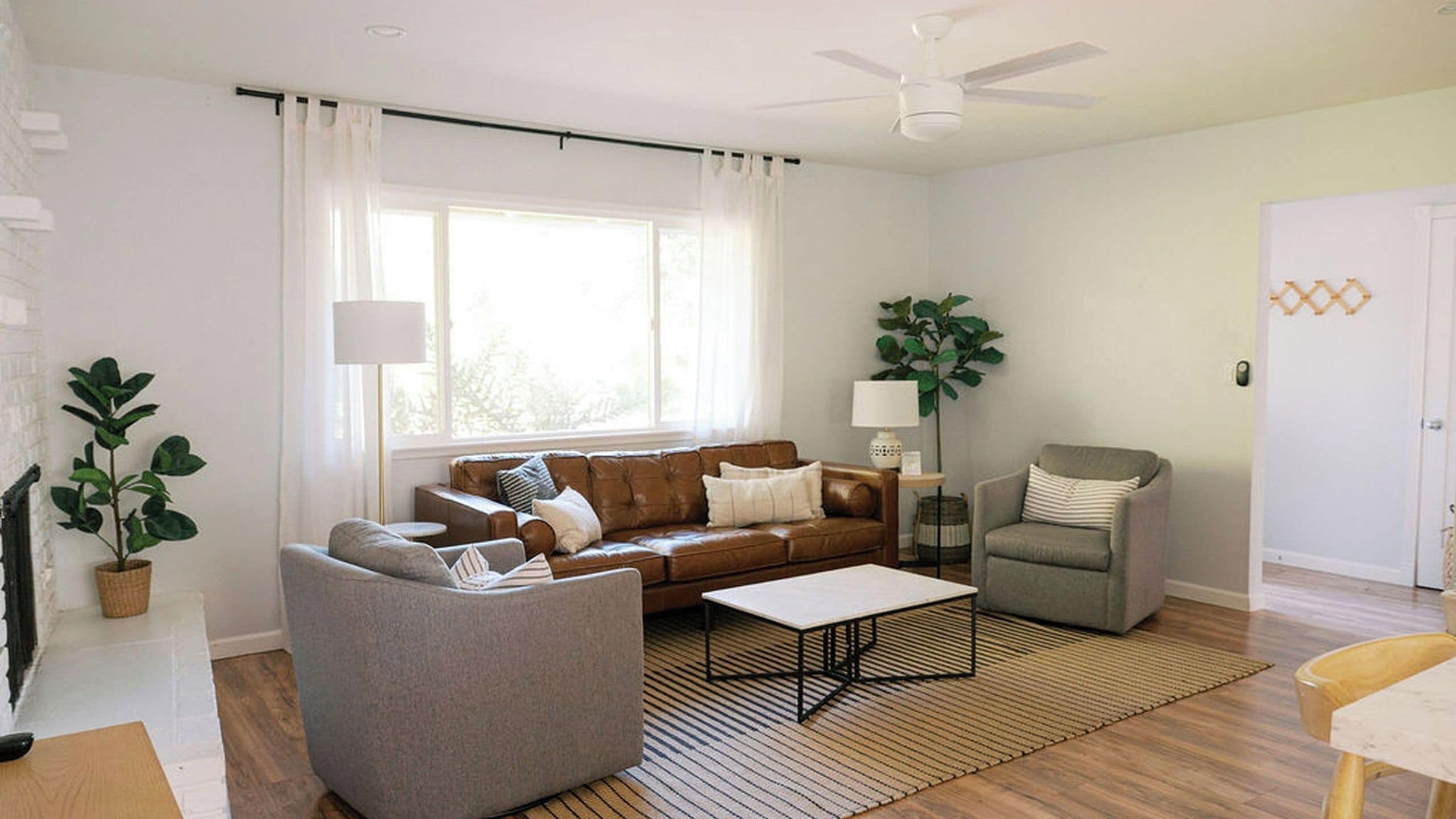 A living room with a brown leather sofa, a grey armchair, a coffee table, and a large window with white curtains.