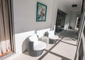 A covered outdoor seating area with wicker chairs and cushions, a wall-mounted bird painting, and a view of the street. -  New Property! The Hidden Catch.. 2-bdr house on the water and great for fishing (Lauderdale-by-the-Sea)