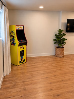 A room with a wooden floor, a Pac-Man arcade machine, a wall-mounted TV, and a potted plant. - Bright 2BR Basement Apt - Near BSU & Micron (Boise)