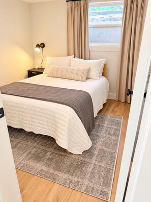 Un dormitorio con cama, una mesita de noche con lámpara, una ventana con cortinas y una alfombra en el suelo.