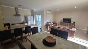 A modern kitchen with a dining area, a living room with a sofa and TV, and a window providing natural light.