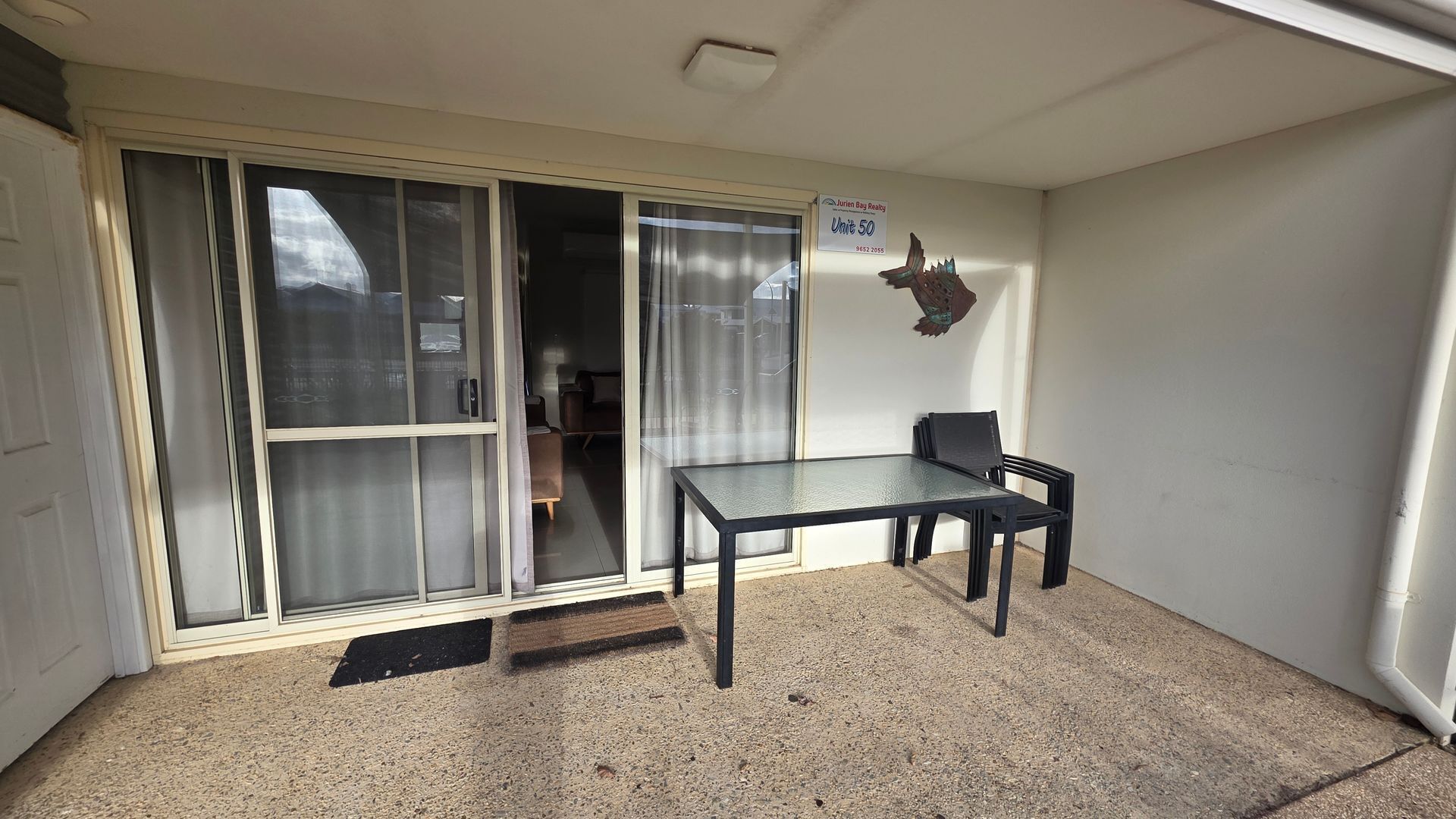 A room with a sliding glass door, a table with two chairs, and a sign indicating 'Jurien Bag Realty Unit 50'.