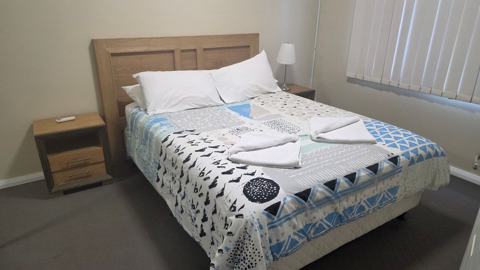 A bed with a patterned bedspread, a wooden headboard, a nightstand with a lamp, and a window with blinds.