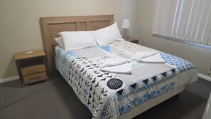 Una cama con colcha estampada, cabecera de madera, mesita de noche con lámpara y ventana con persianas.